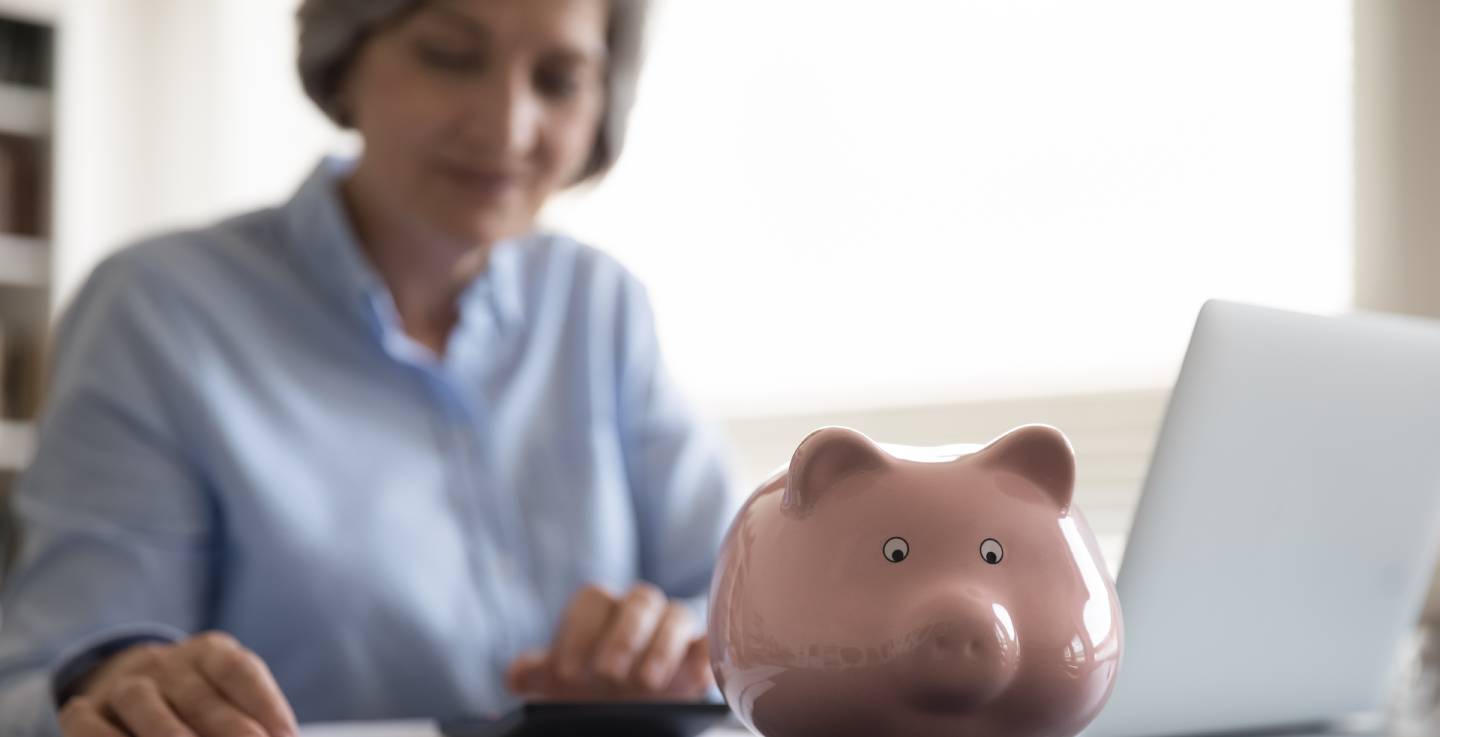 Classic piggy bank on desk, older woman sits at a desk with laptop using a calculator 