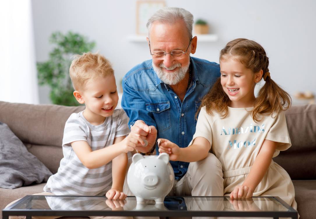 Aged man giving piggy bank to boy and girl while showing grandchildren how to save money at home