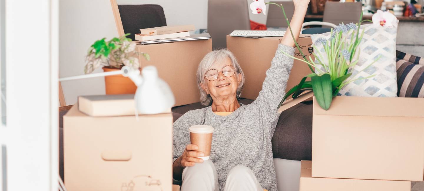 Happy senior woman involved in moving house sitting among cardboard boxes takes a break with a coffee, concept of moving, retirement, new life, buying, renting, apartment, house