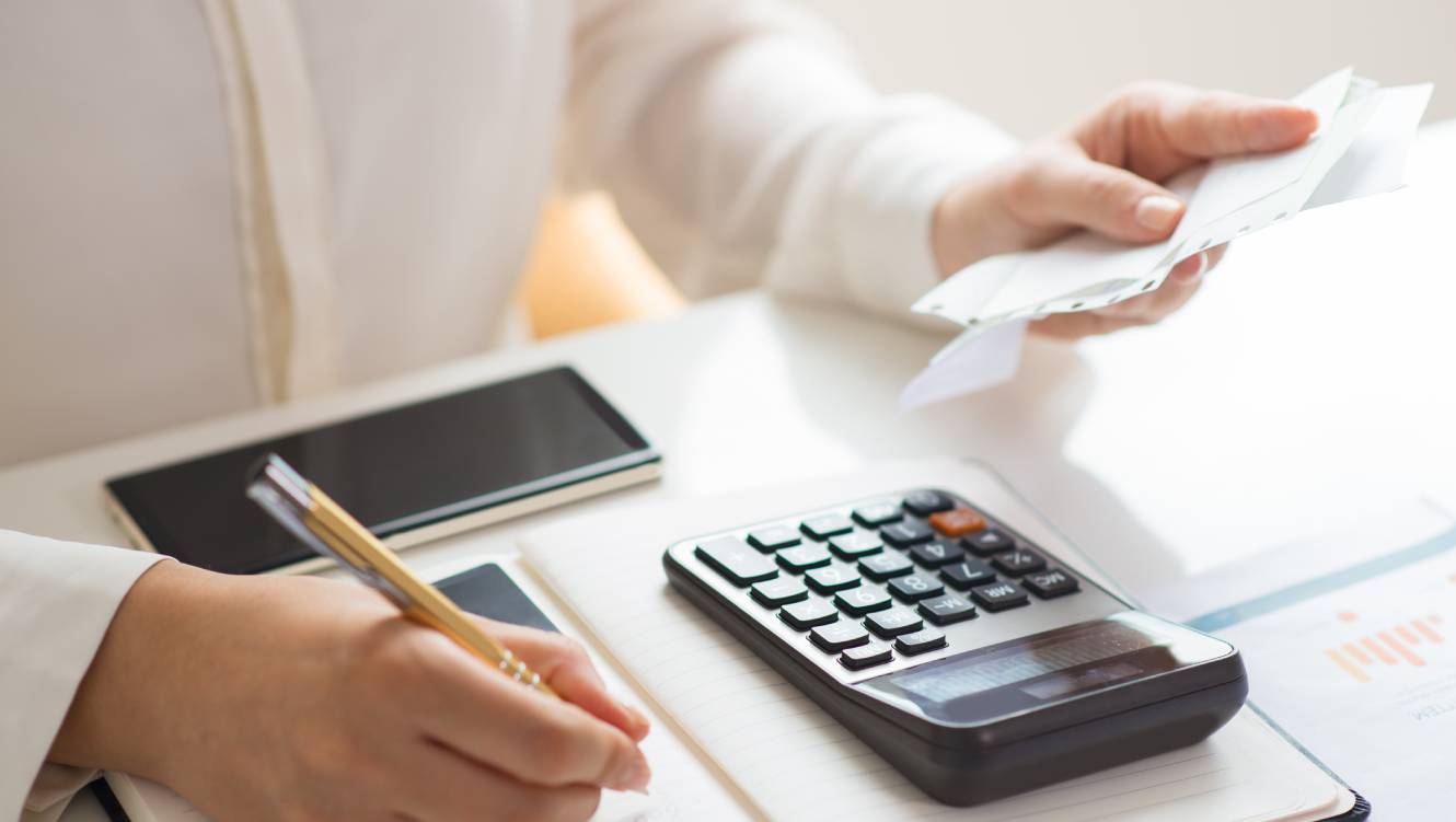 Closeup of person holding bills and calculating them. Notebook, calculator and smartphone lying on desk. 