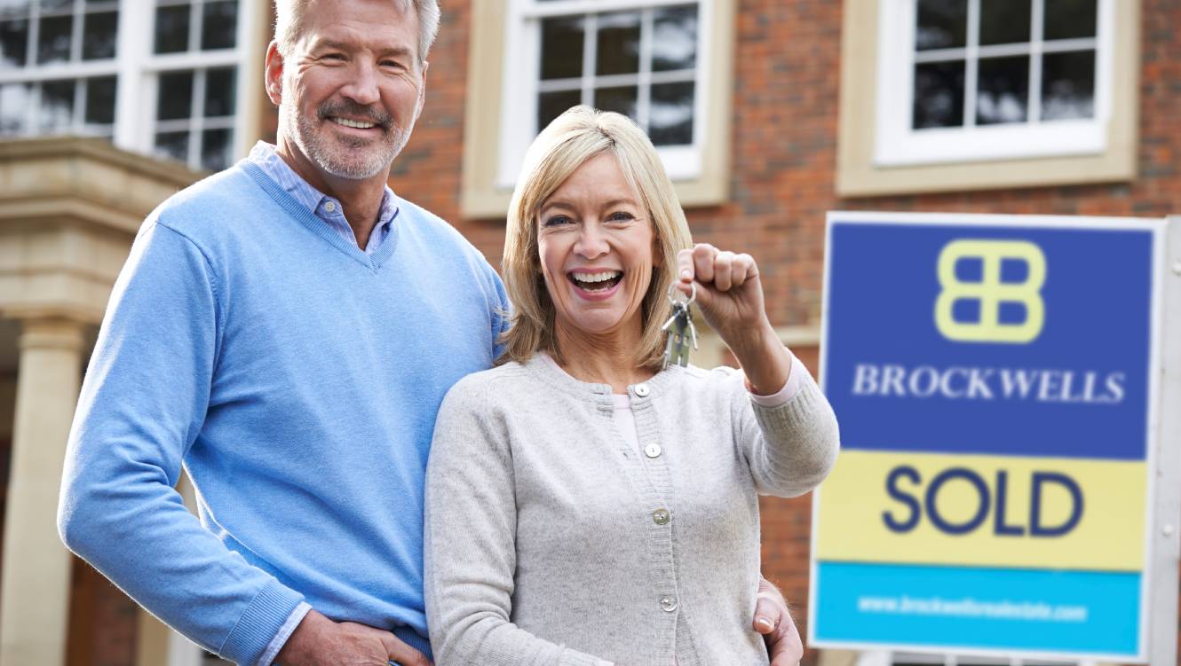 Mature Couple Holding Keys To New Home Standing By Sold Sign