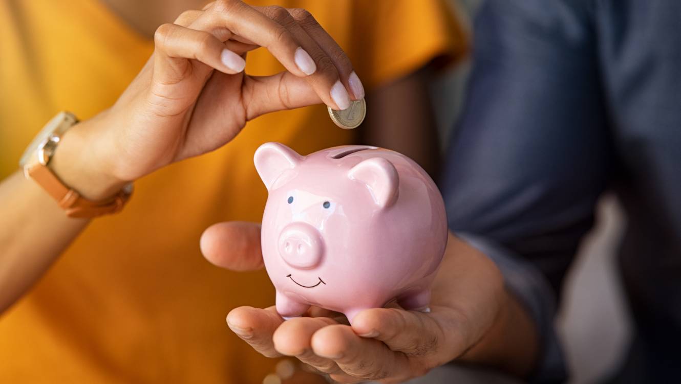 Close up of man holding pink piggybank while woman putting coin in it. Indian young couple saving money for their wedding. Close up of woman hand putting money in piggy bank to save.