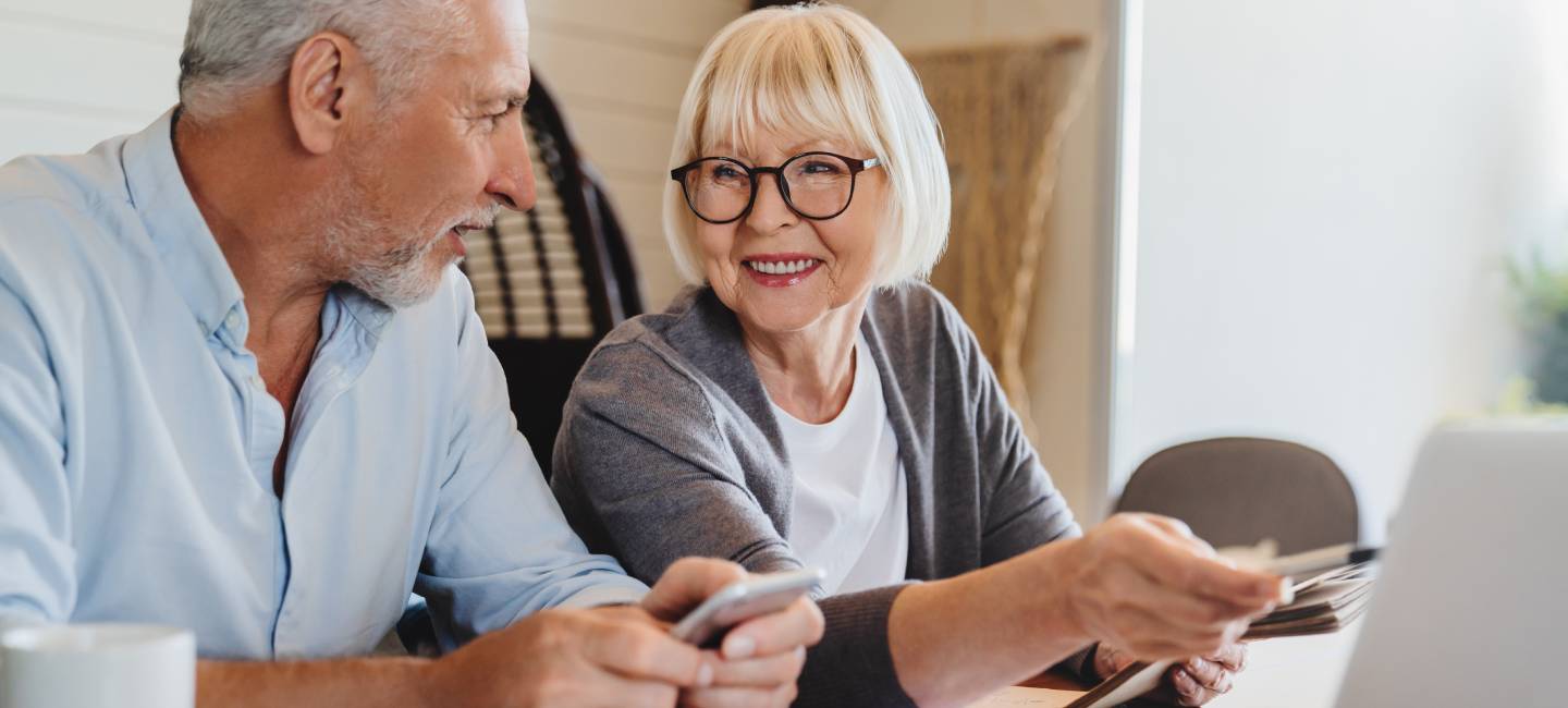 Cheerful elderly grey haired wife and husband sit on couch using computer