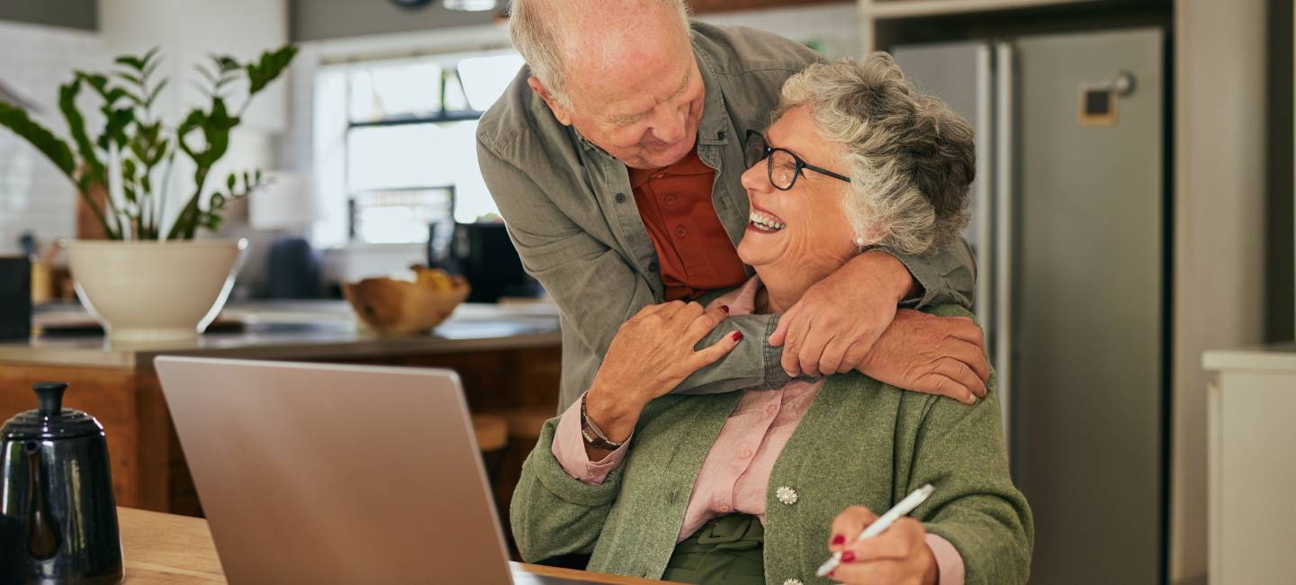 Elderly couple collaborating on financial home on laptop at home while sharing an affectionate moment.