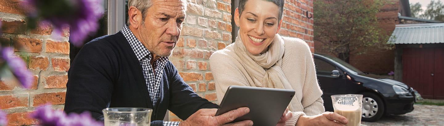 A mature couple are looking intently at a tablet computer as they drink coffee at a courtyard table