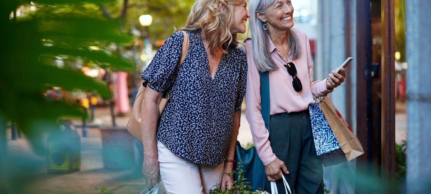 Two older women happily looking at a shop window during shopping time together.
