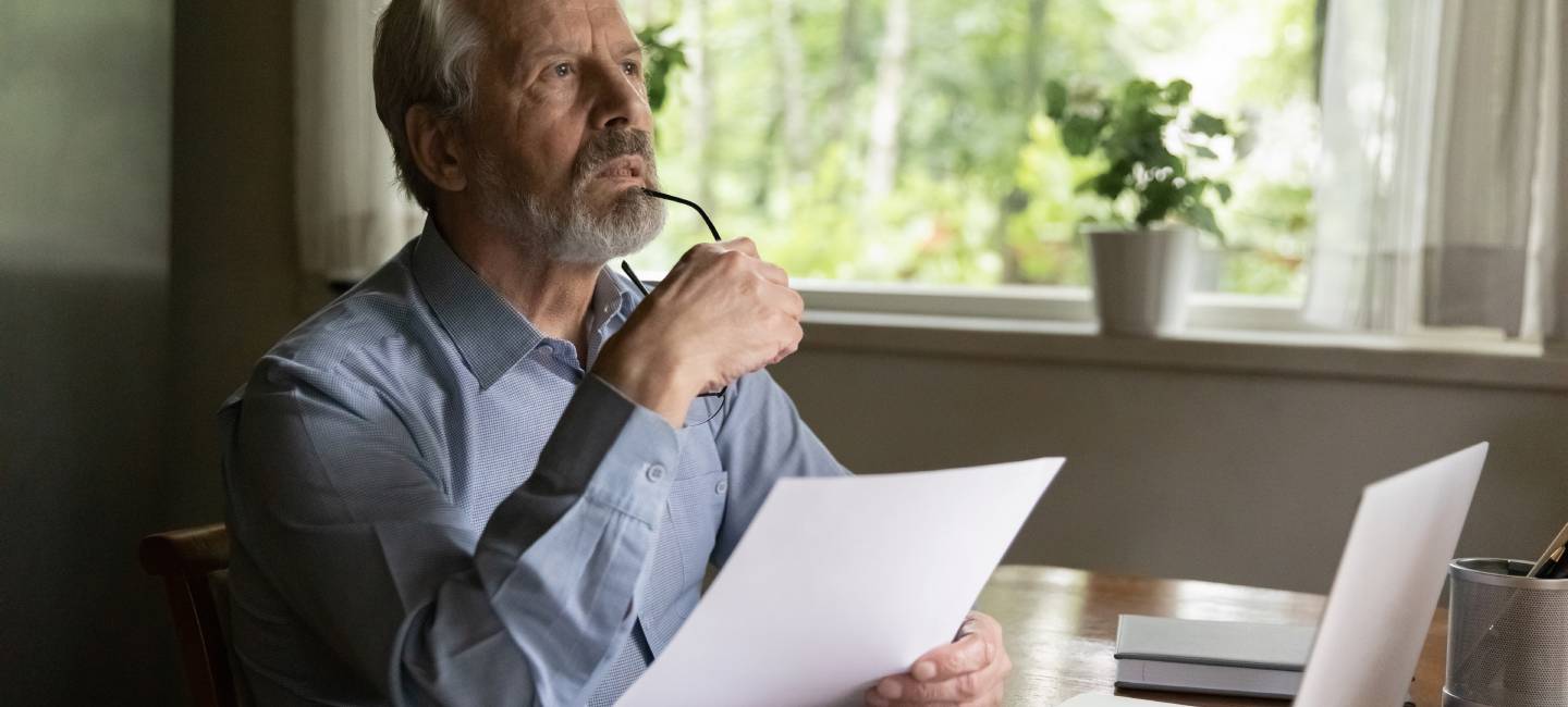 Thoughtful aged man ponder on document at office table before laptop 