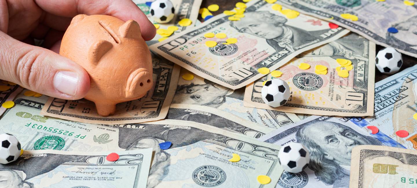 Hand placing a piggy bank on dollar bills with small soccer balls and tricolor confetti, concept of investment or soccer winnings.