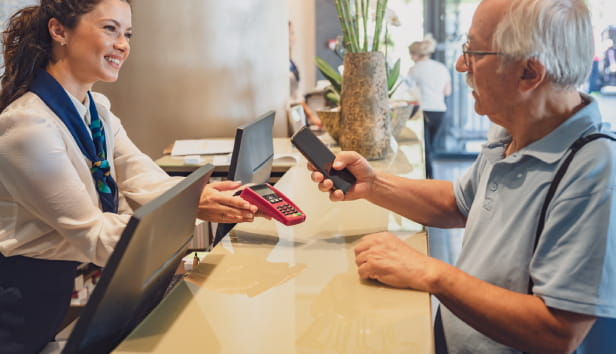 A senior couple at the hotel front desk makes contactless payments with a smartphone. Seniors traveling and using new technologies.
