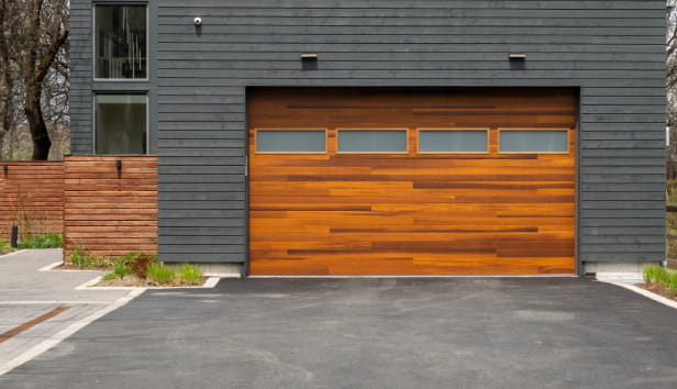 A modern brown faux wooden exterior garage door with four small horizontal glass windows. The modern door is on a luxury dark grey contemporary house with a concrete driveway. 