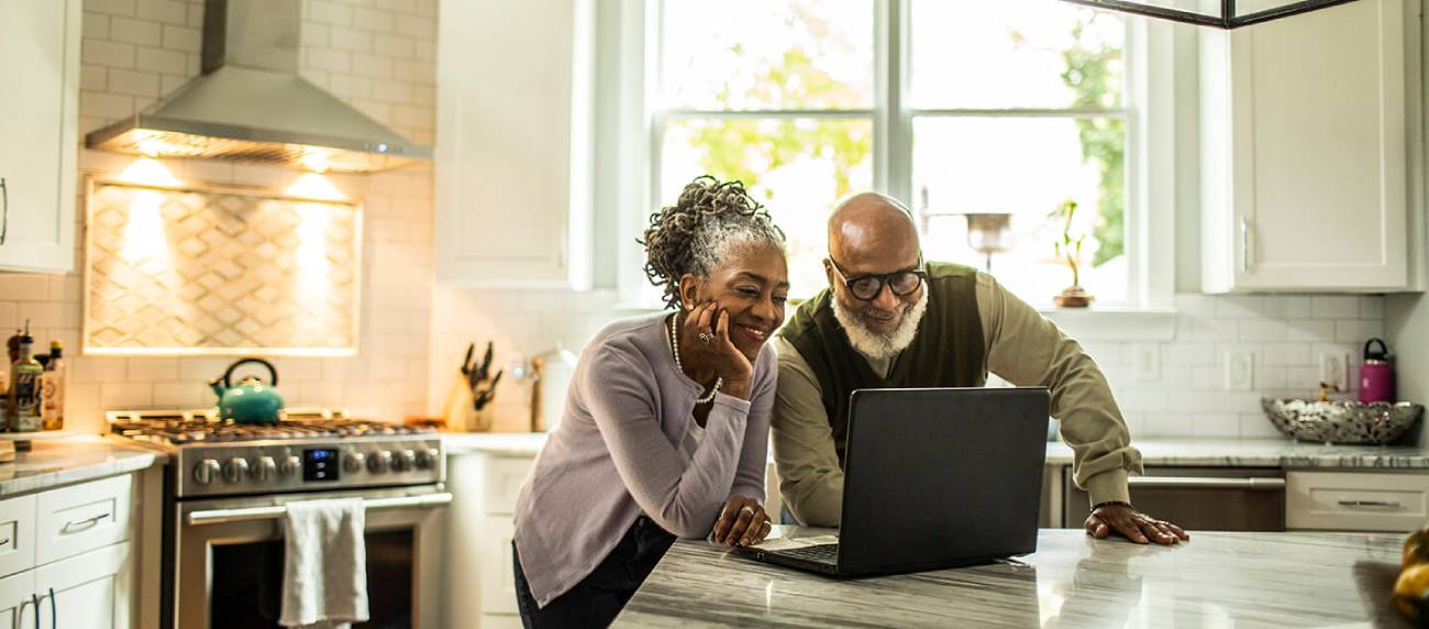 Mature couple standing in their kitchen typing on their laptop