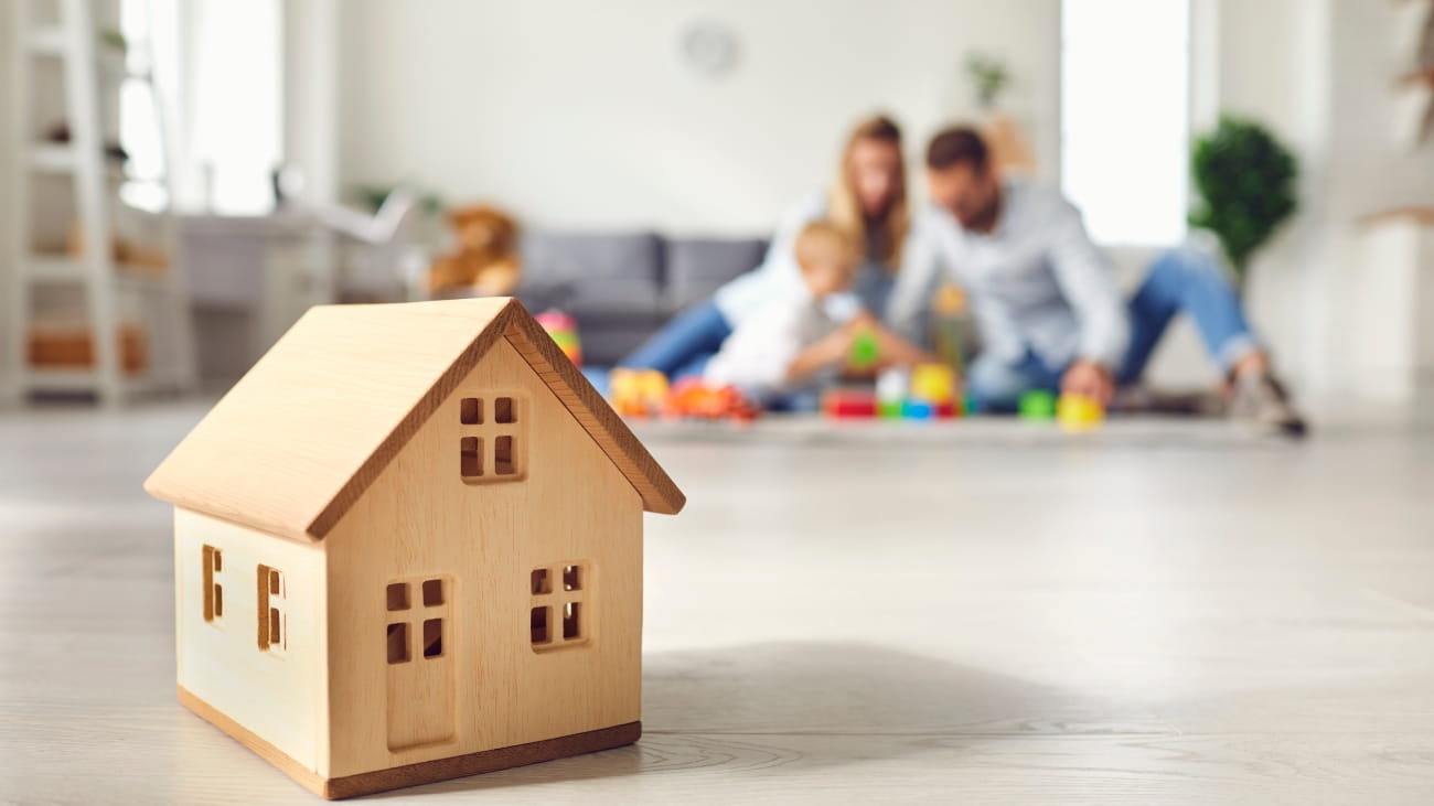 A mini house made from wood. In the background is a a mother and father playing with their child 