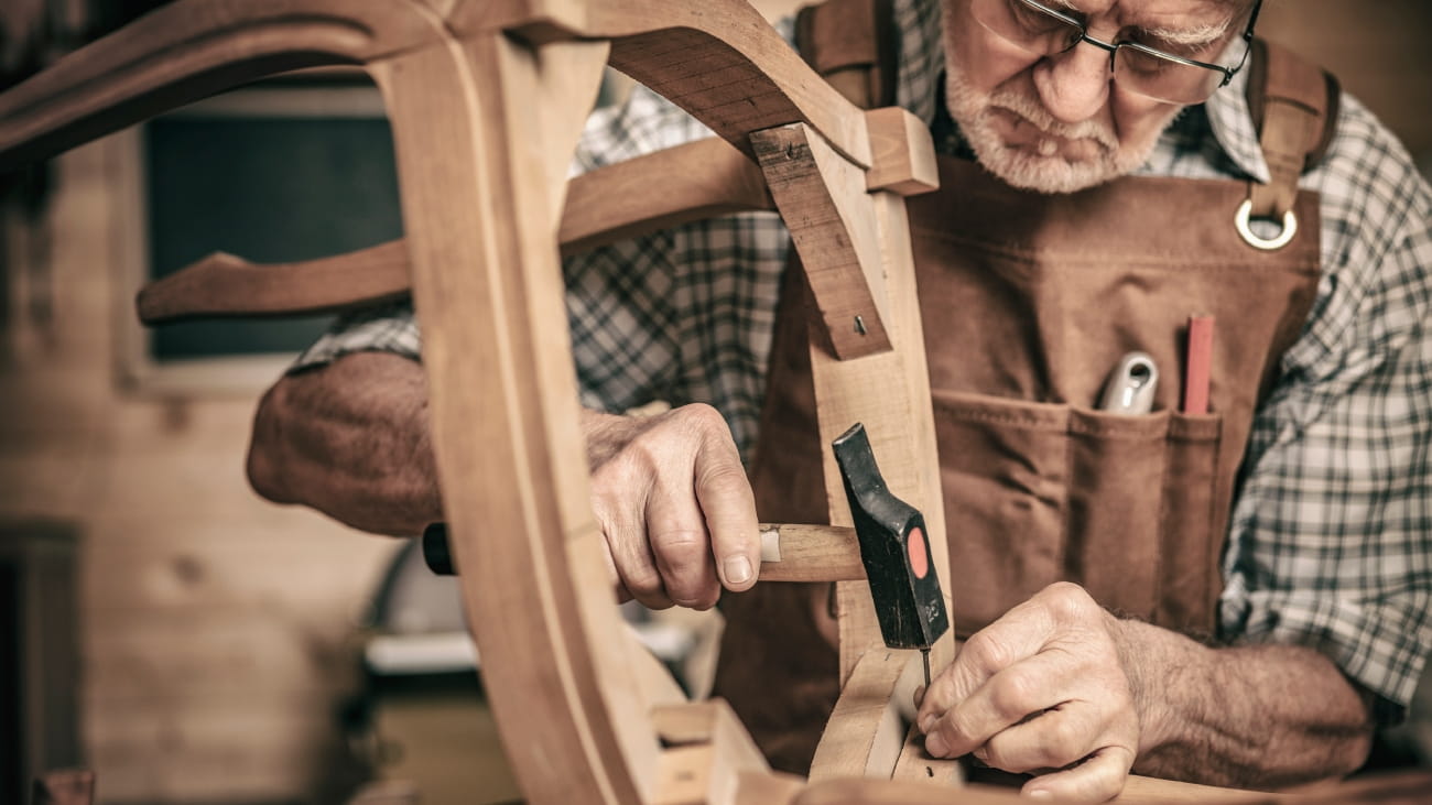 An older man putting a nail in to a chair using a hammer
