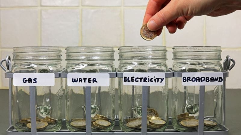 Four mason jars filled with pound coins are lined up. Each has a label on them - Gas, Water, Electricity and Broadband. A person is adding another coin to a jar.