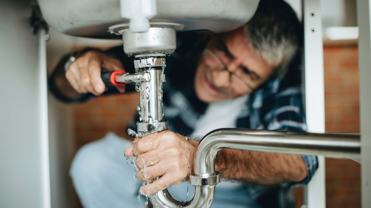 A man crouching under a sink trying to fix  the pipe