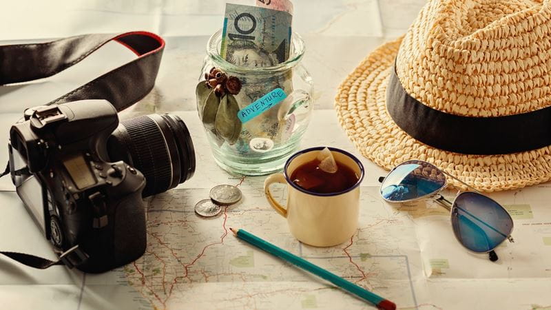 A table with lots of exciting items indicating someone is on holiday - a camera, sunglasses, a sunhat, a map and foreign currency in a jar that says 'Adventure' 