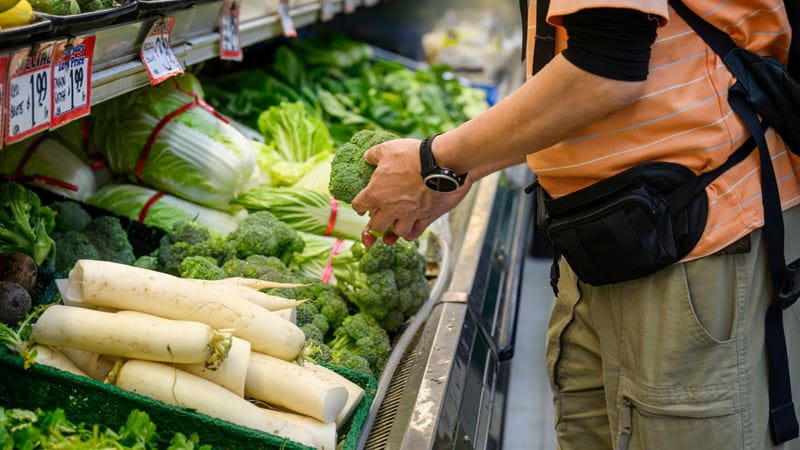 A backpacker wearing a travel money belt, choosing fresh vegetables at the supermarket. Customers buying food at grocery shops. USA.