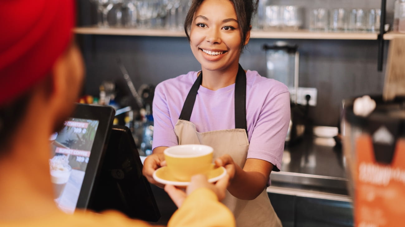 Smiling barista handing a cup of coffee to a customer in a cafe. The barista is wearing a purple shirt and a beige apron