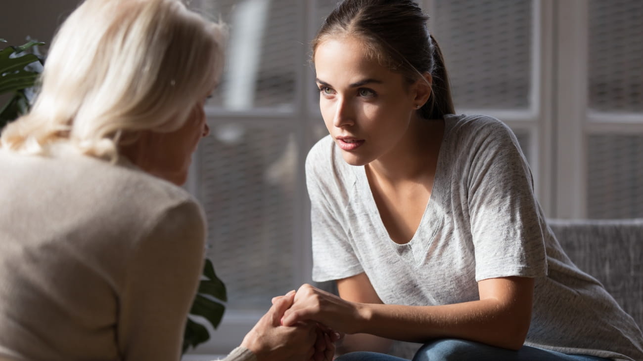 Grown up daughter holds hands of mid aged mother sit on armchair looking at each other having heart-to-heart talk, concept of close relative person, diverse generations connection, mom and adult child.