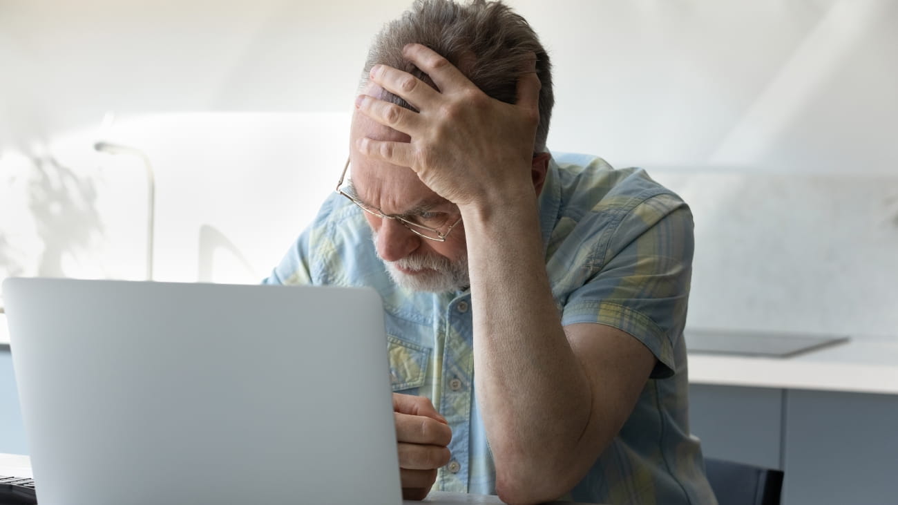 Frustrated tired senior man touching head at laptop, receiving shocking bad news, siffering from headache, migraine, memory loss, losing money due to fraud, scam, insecure financial data, hacking.