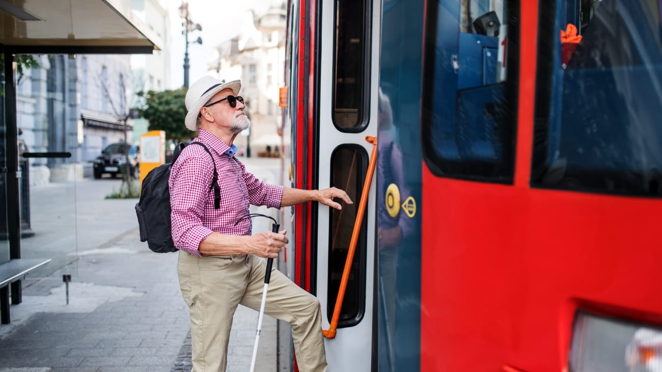Senior blind man with white cane getting on public transport in city.