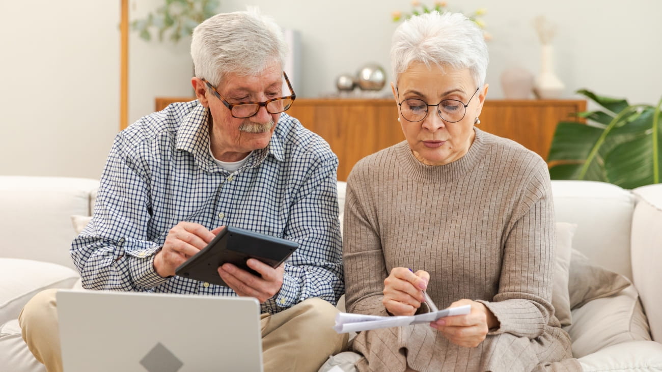 Mature couple sit with laptop and paper document calculating pension savings.