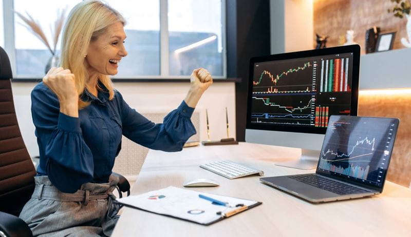 Happy joyful mature blonde business woman, trade broker, stock trader, sits at table in office, shouts and rejoices at success, good profit, looks at charts on pc and laptop, gestures with fists