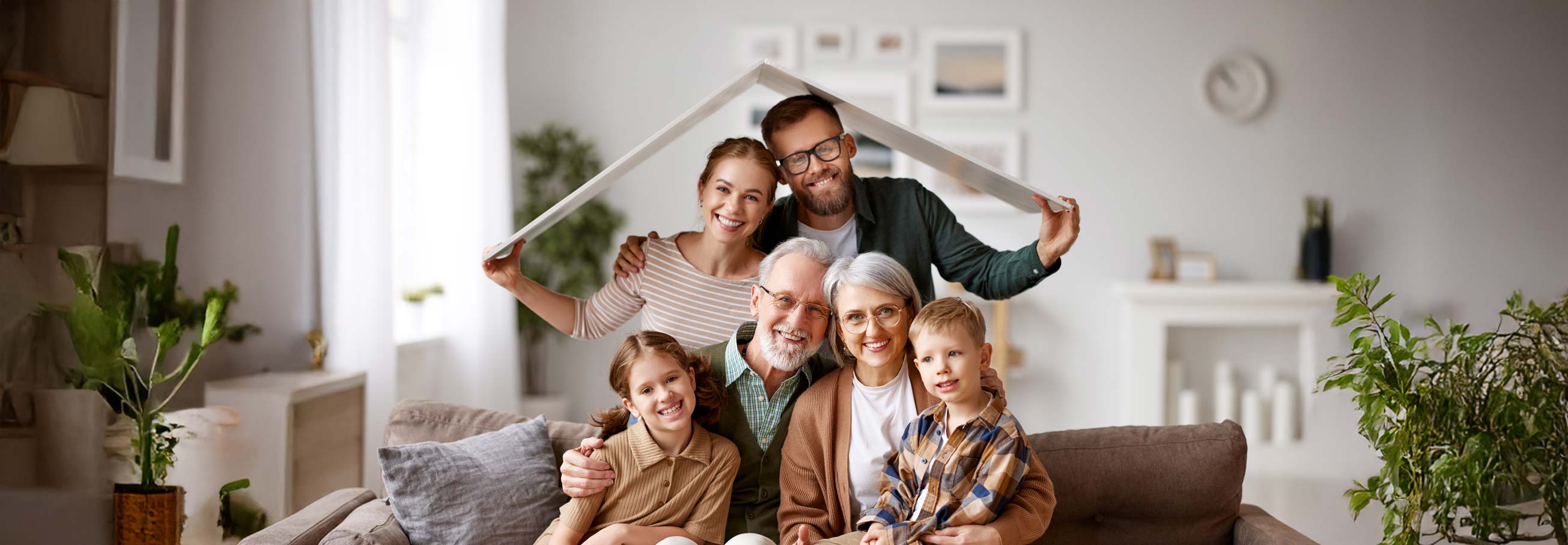 Big happy family, grandparents, mother, father with little kids son and daughter celebrating relocation in new home, sitting on the coach under paper roof and smiling at camera. Mortgage loan concept