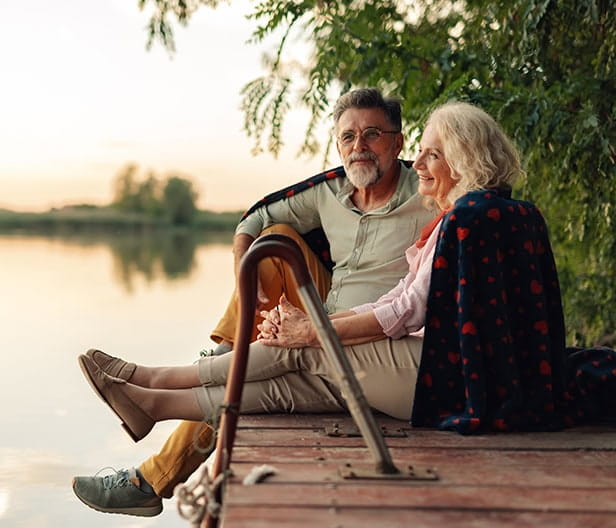 Mature couple relaxing on a pier, sharing a blanket and enjoying the sunset over the lake.